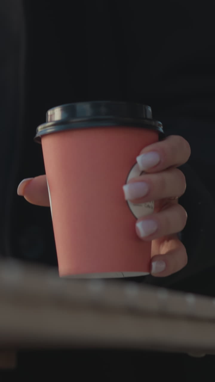 primer plano de alguien con uñas pulidas colocando una taza de café en la mesa frente a una computadora portátil, fondo borroso con estructura poco clara, persona a punto de sentarse en una mesa al aire libre