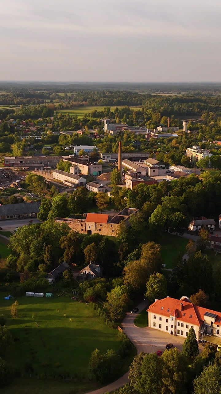 Aerial ascend of Aizpute, Latvia, showing a historic town surrounded by greenery and countryside, vertical at sunset