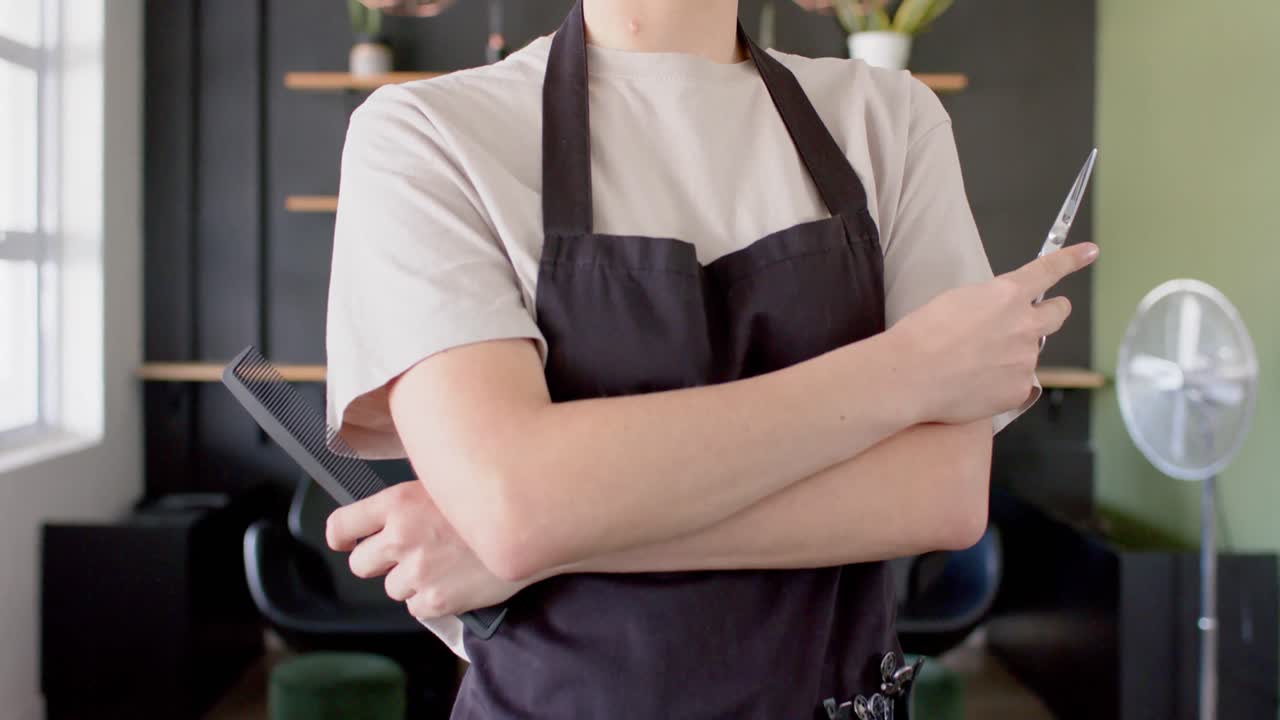 Midsection of caucasian female hairdresser in black apron holding scissors and comb, in slow motion