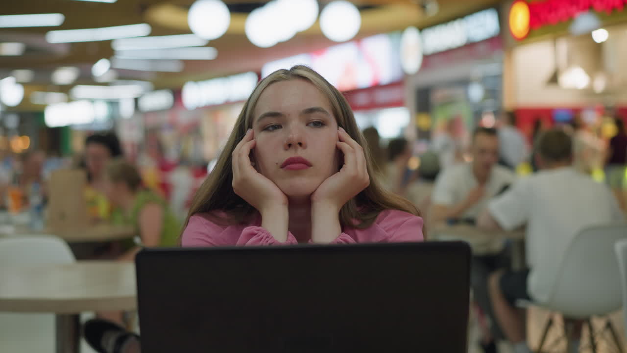Young woman in pink dress sits in front of her laptop, resting her chin on both hands and appearing deep in thought, blurred background of people in a busy, well-lit restaurant, with blur light