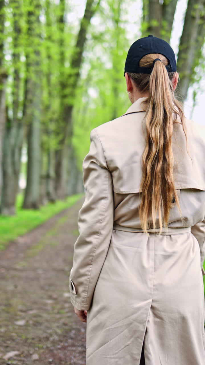 Una bella giovane donna in impermeabile passeggia lungo un sentiero in un parco, vicino agli alberi, in un ambiente tranquillo nel primo pomeriggio. Una donna graziosa a passeggio in un parco all'inizio della primavera.