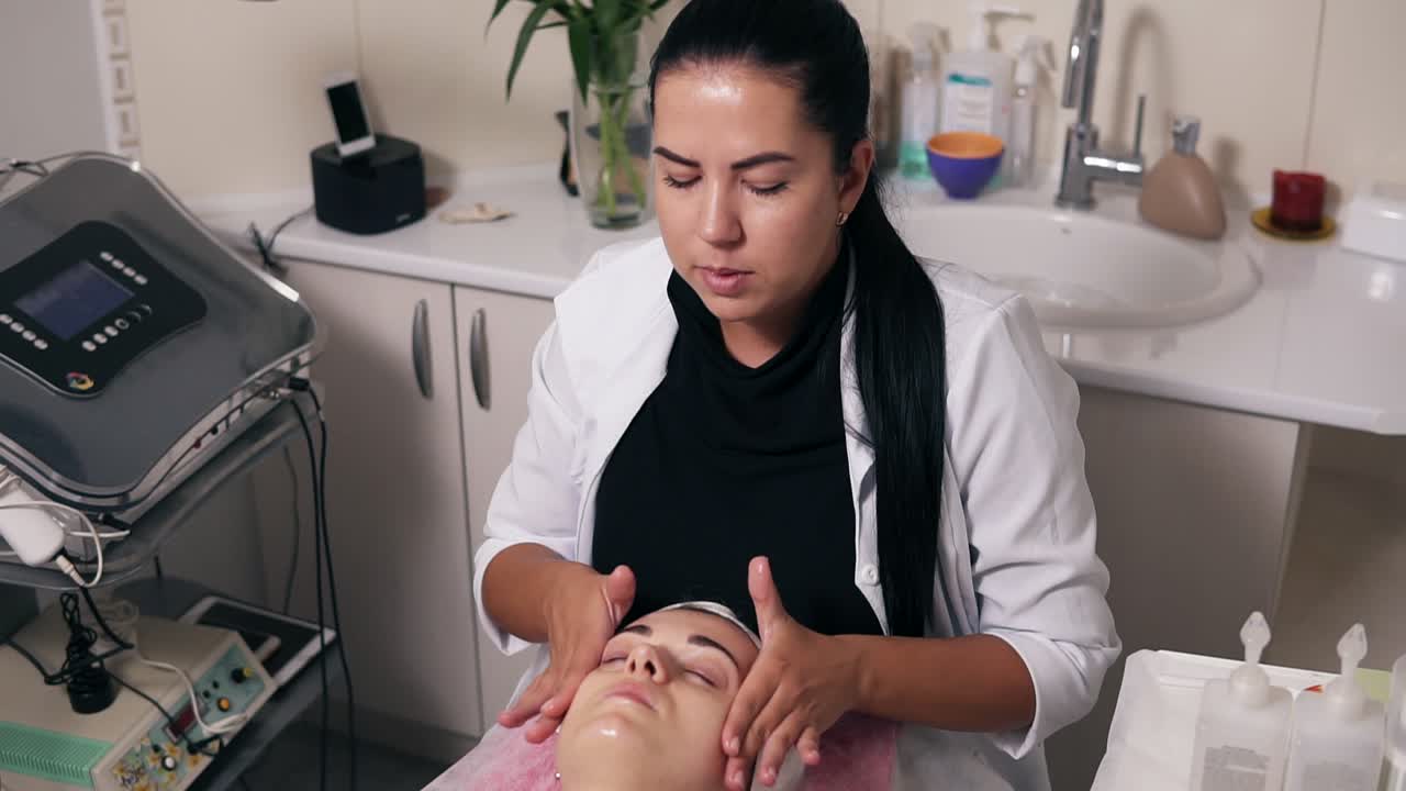 Female cosmetologist is making face massage in spa salon. Young woman with her eyes closed is lying on the couch during cosmetic