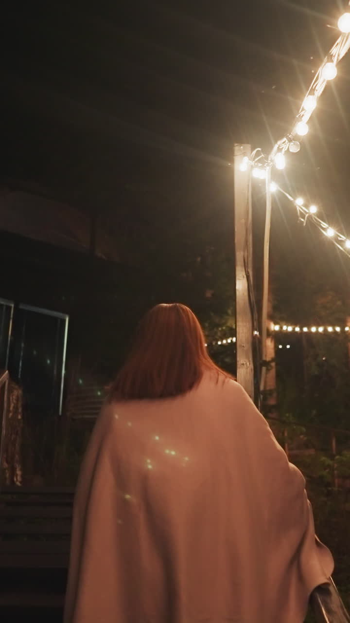 una mujer joven con una manta sube las escaleras. una mujer solitaria regresa a casa de la caminata de la noche a una casa de madera. la escalera iluminada con bombillas decorativas