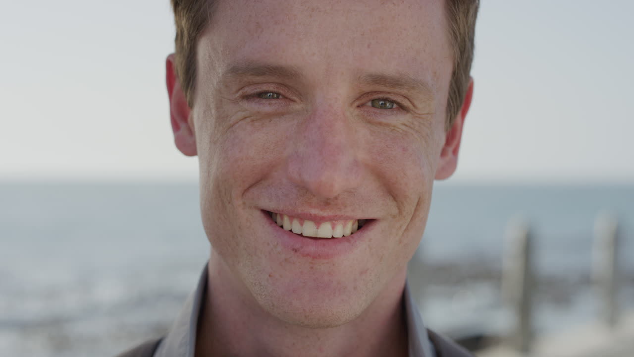 retrato de cerca joven apuesto de cabeza roja sonriendo estudiante feliz disfrutando de un estilo de vida exitoso relajándose en un cálido día de verano en la orilla del mar cámara lenta