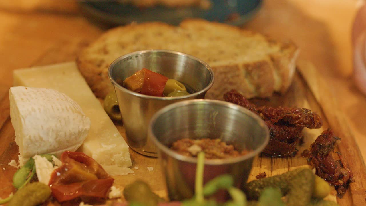 A fork lifts an olive from a metal cup on a rustic antipasto platter with assorted cheeses, pickles, bread, and sun-dried tomatoes under warm lighting