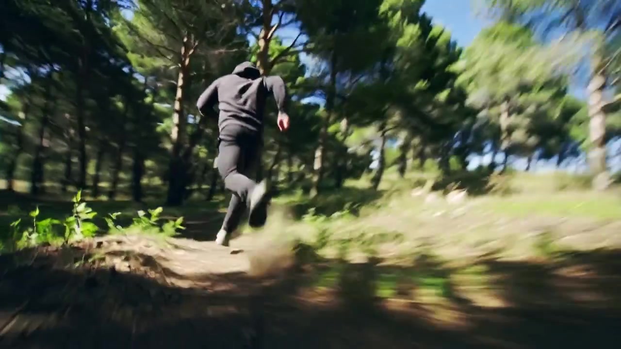 Camera starting behind hooded man in pine woods, filming him running, dodging trunks, dust rising