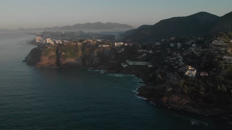 Luftaufnahme-Der-Klippen-Von-Joatinga-Beach-In-Rio-De-Janeiro-Mit-Im-Hintergrund-Der-Riesigen-Nachbarschaft-Von-Barra-Da-Tijuca-Mit-Morgennebel-Bei-Sonnenaufgang