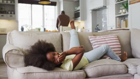 hija afroamericana acostada en el sofá riendo en la televisión con el padre en el fondo