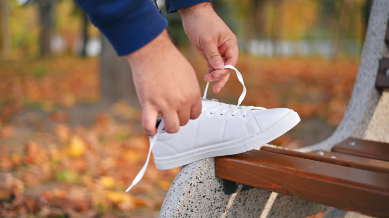 Slow motion view of a man with prosthetic legs and white sneakers. Tying shoelaces with his foot on the bench in a park. Fallen yellow leaves around
