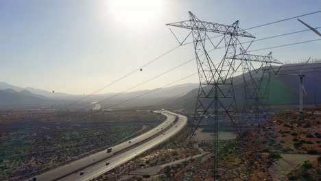 Aerial-view-panning-from-right-to-left-alongside-wind-turbines-and-high-voltage-transmission-lines-revealing-highway-and-mountains-near-Palm-Springs-in-the-Mojave-Desert,-California,-USA