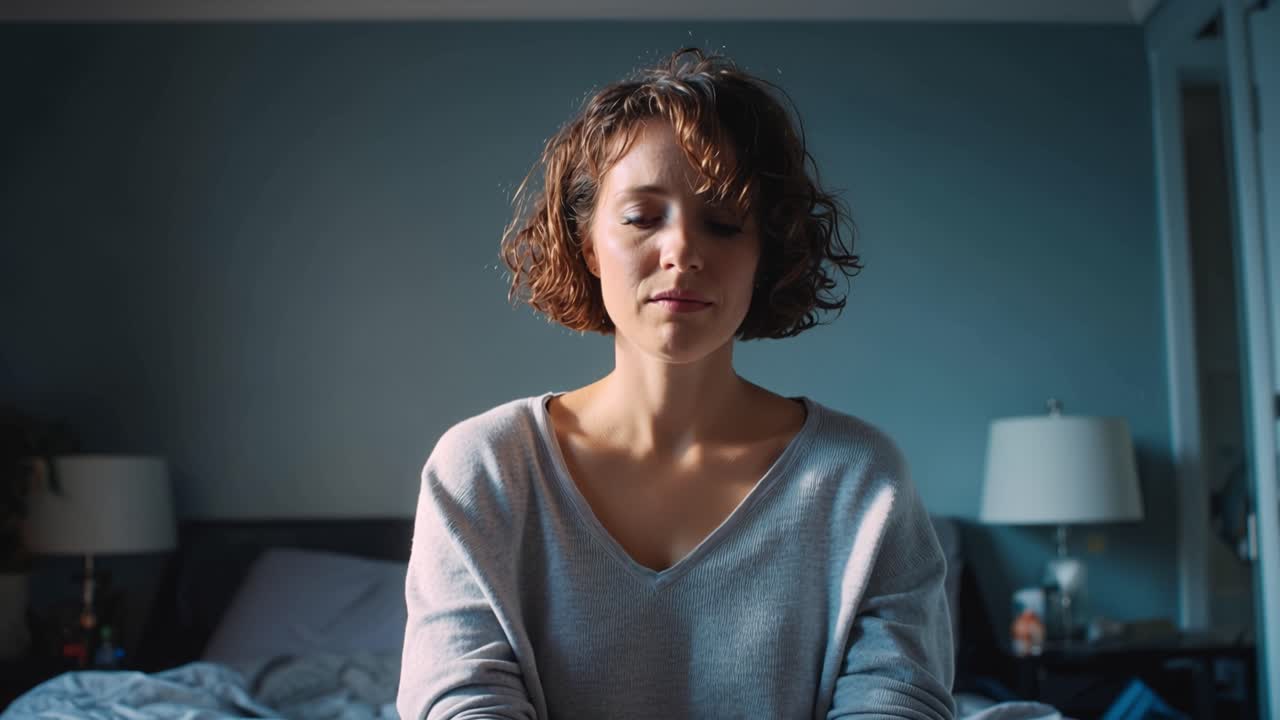 Woman sits on bed, head in hands. She appears stressed or tired. She slowly lifts her head, then stretches. Her face shows quiet discomfort or fatigue