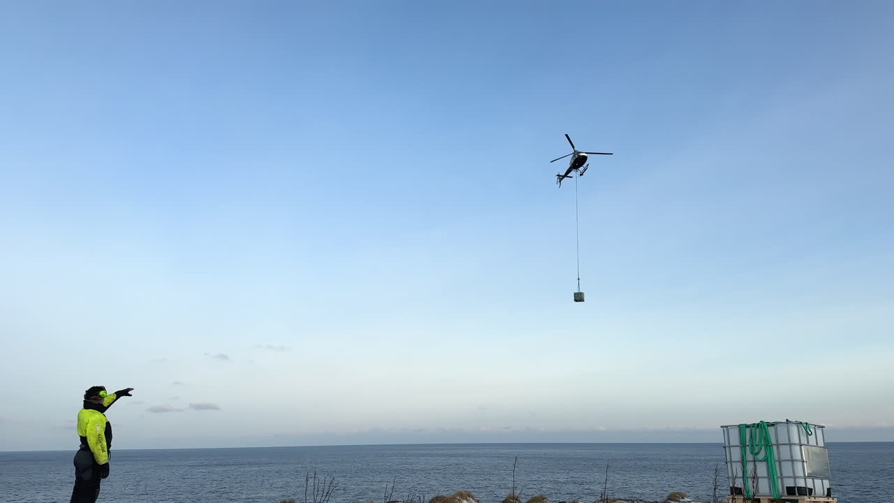 Shot of a helicopter carrying cargo on a longline to a remote island off the coast of Norway with a spotter directing
