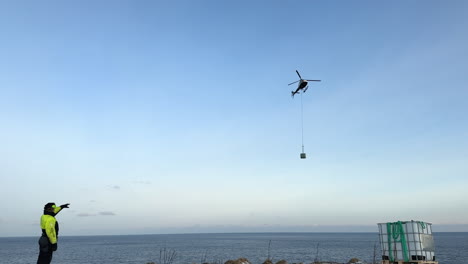 shot of a helicopter carrying cargo on a longline to a remote island off the coast of norway with a spotter directing