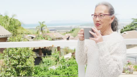 happy asian senior woman relaxing on balcony with mug of coffee