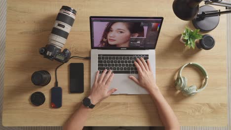 top view zoom out shot of a male editor sitting in the workspace using a laptop next to the camera editing photo of a female at home