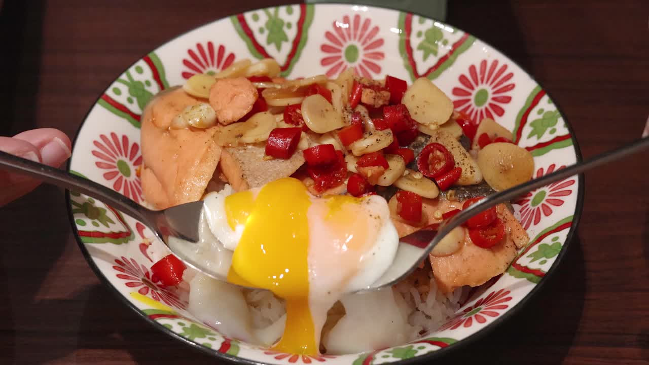 A spoon and fork break and mix a soft-boiled egg over rice, salmon, garlic, and chili in a patterned bowl under warm indoor lighting