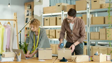 Hombre-Caucásico-Que-Trabaja-En-Una-Tienda-De-Ropa-Empacando-Pedidos-En-Paquetes-Mientras-Una-Mujer-Caucásica-Escribe-En-Una-Computadora-Portátil-En-Línea