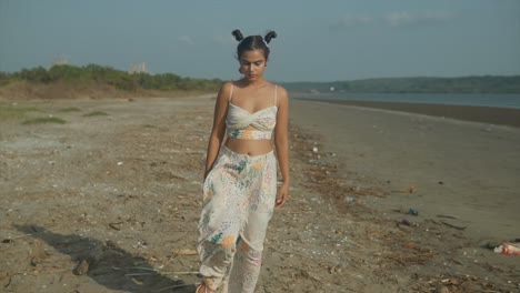 cinematic shot of an indian fashion model wearing a white and colored trousers and top walking on a sandy beach on a wind and sunny day in goa, india