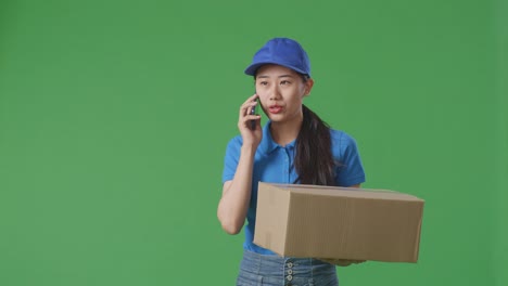asian female courier in blue uniform walking and talking on smartphone while delivering a carton on green screen background in the studio