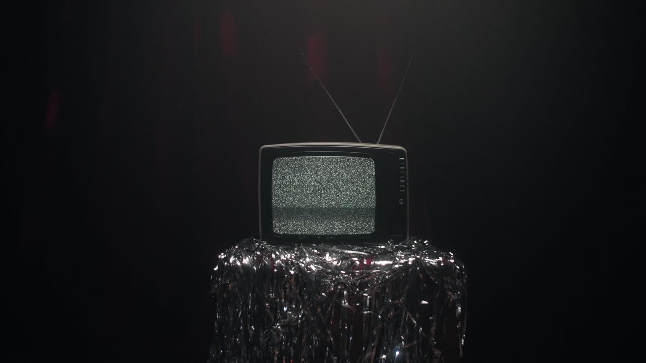 Beyaz gürültü ve analog parazit gösteren eski bir vintage televizyonun yakın çekimi. Sinematik kamera yakınlaştırma ve uzaklaştırma hareketi. TV, koyu ve kasvetli bir arka plana karşı parlak alüminyum folyo üzerinde duruyor