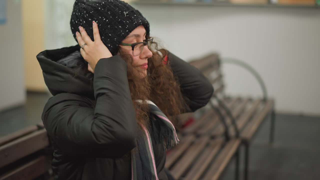 Girl in black jacket sitting on bench adjusts long curly hair with hand, head bent forward, showing detail of polished nails and casual winter style in candid indoor lifestyle moment with relaxed expression