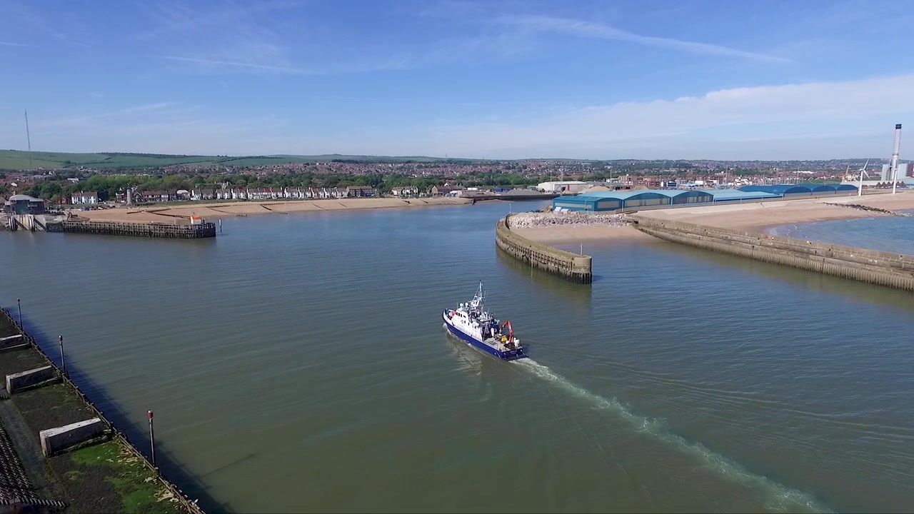Ripresa con drone che segue un peschereccio che entra in un porto con una città costiera e colline ondulate sullo sfondo