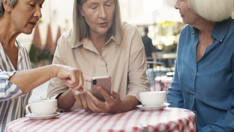 Drei-ältere-Frauen,-Die-Freundlich-Auf-Das-Smartphone-Schauen-Und-Im-Sommer-Kaffee-Am-Tisch-Auf-Der-Caféterrasse-Trinken