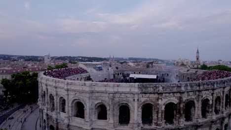 Drone-over-the-Arena-of-Nimes-at-sunset,-people-are-waiting-for-the-stromae-concert,-foreground-of-trees