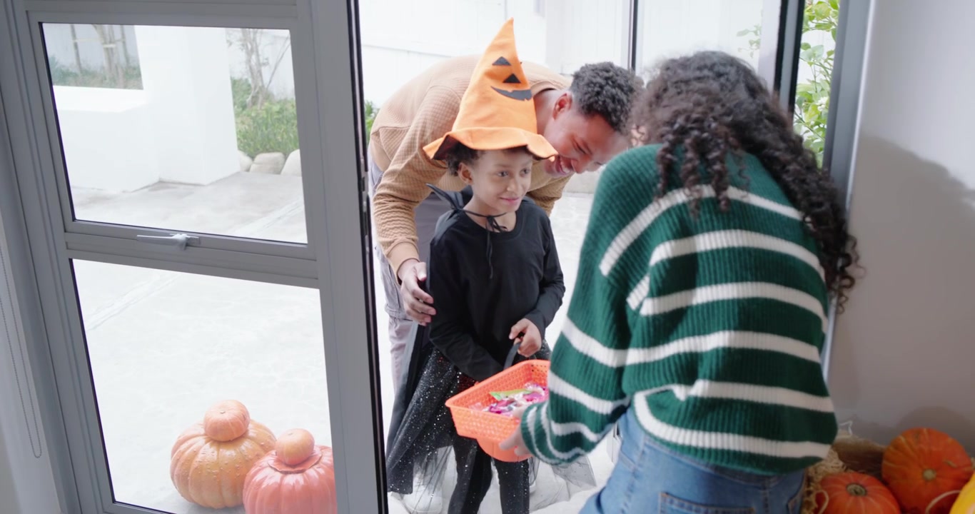 Afroamerikansk barn i græskarhat når døren, mens en kvinde rækker en kurv til Halloween