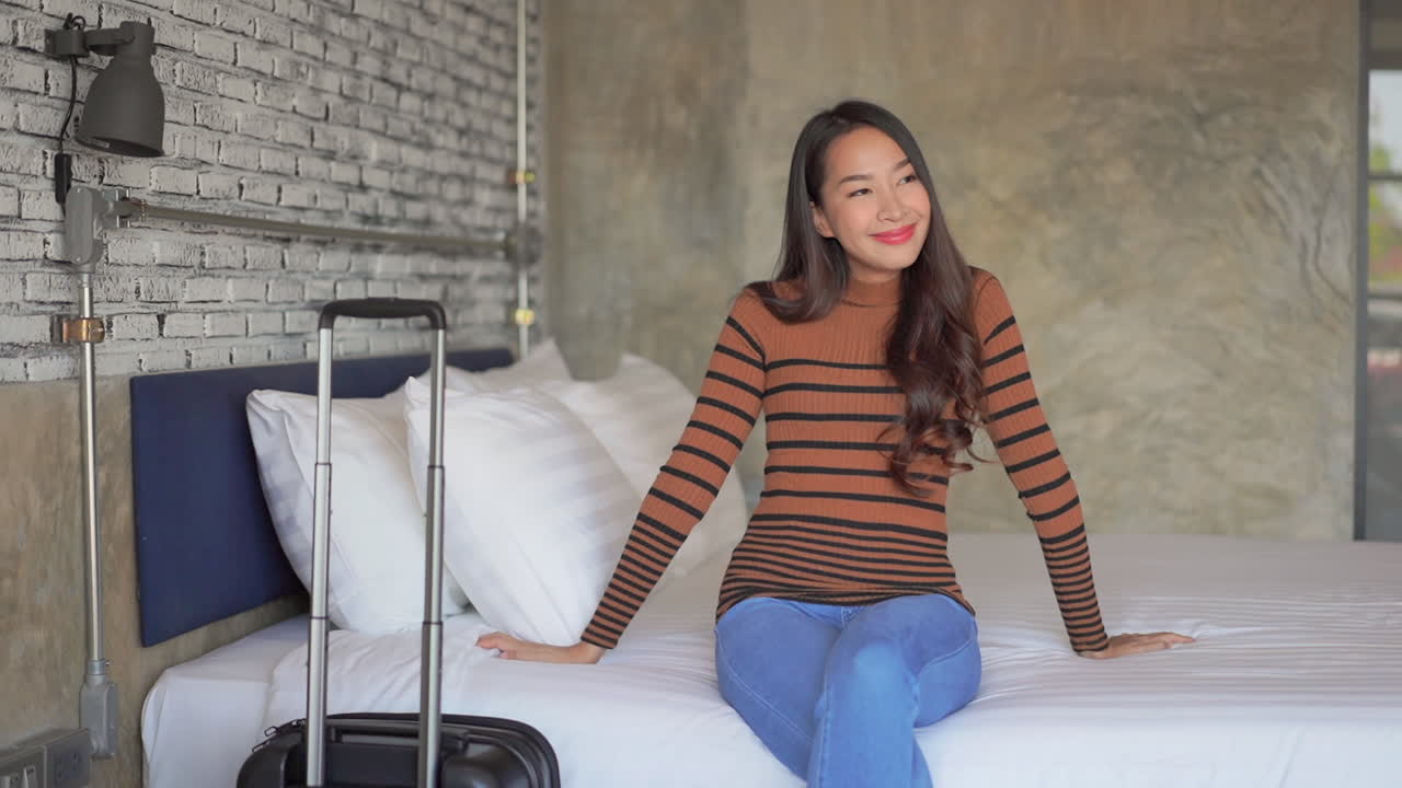 Happy female traveler arriving to her bedroom after checking-in to a hotel, and sitting on her bed with carry-on luggage near the bed