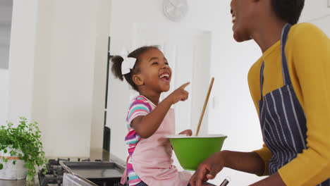 Happy-african-american-mother-and-daughter-wearing-aprons-having-fun-while-cooking-in-kitchen