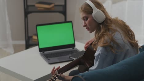 pretty female musician is playing guitar in apartment, green screen of laptop on table in room, online streaming