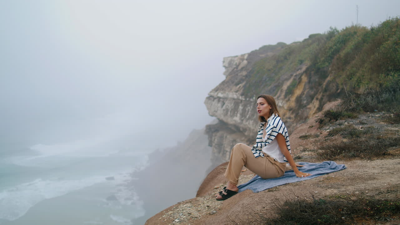 Turista seduta sul bordo di una scogliera con le onde dell'oceano, formato verticale. Ragazza attraente che contempla la vita.