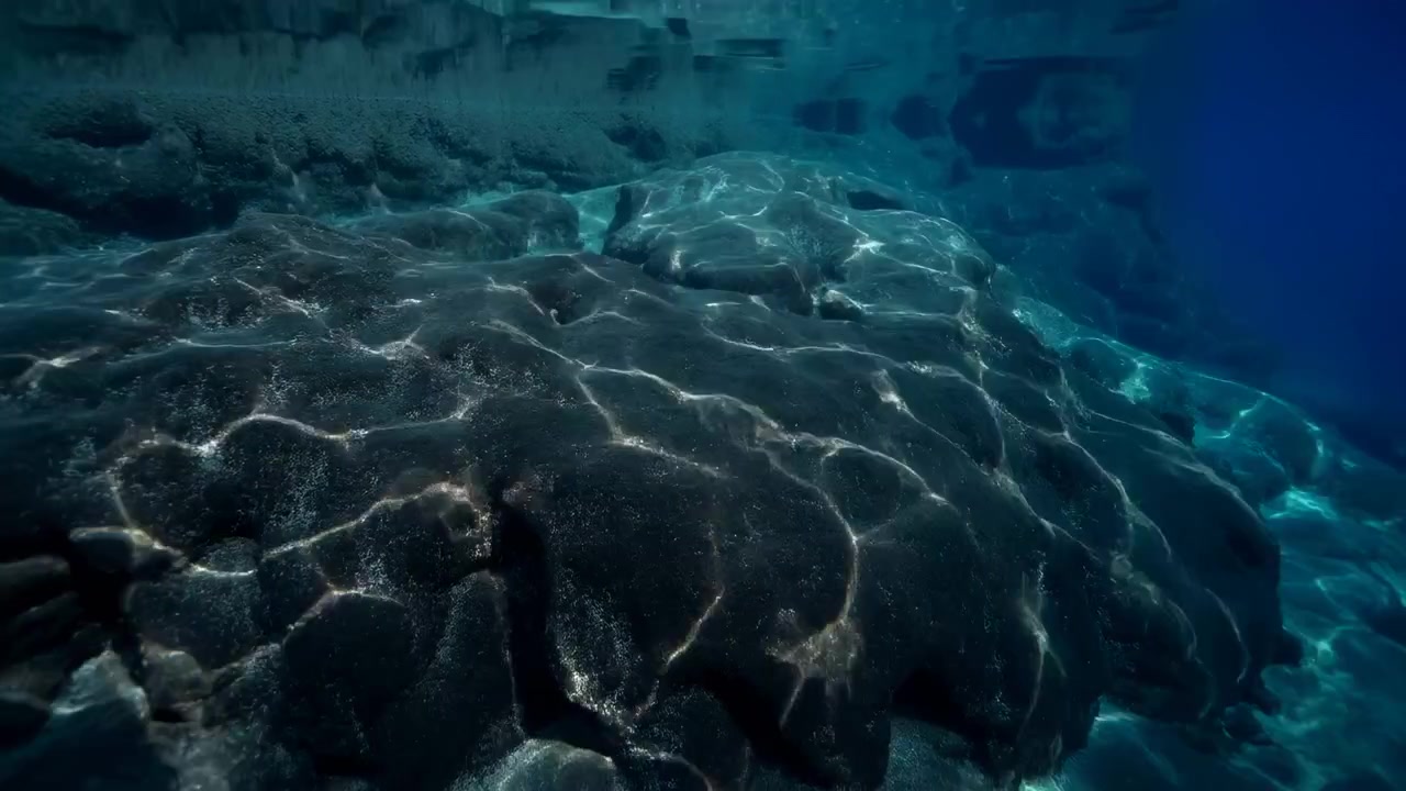 Panoramic underwater scene of dark volcanic rock plateau filling foreground and midground left-to-center, sloping toward deeper blue water at right