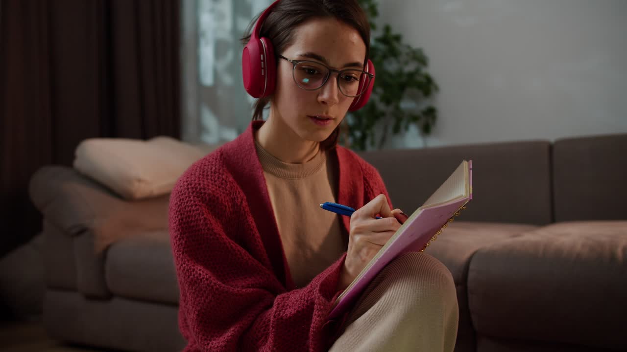 primer plano de una chica morena segura con gafas en un suéter rojo y auriculares inalámbricos estudiando idiomas extranjeros en línea y tomando notas en su cuaderno en un apartamento moderno