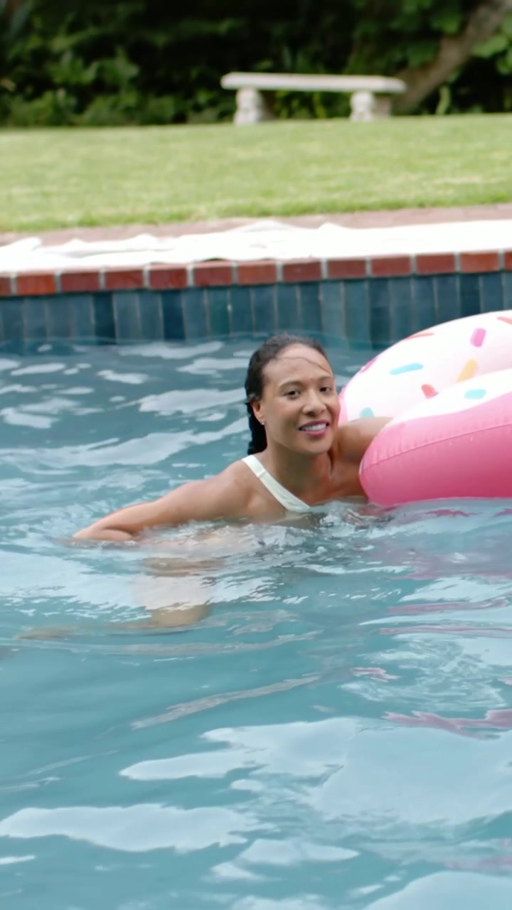 Vertical video: Spotting pink donut, woman in white suit swimming in pool gripping float, climbing