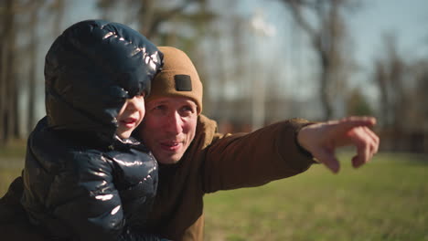 father squatting and holding his son on his leg, the father is smiling and pointing excitedly at something in the distance, the father is wearing a head warmer