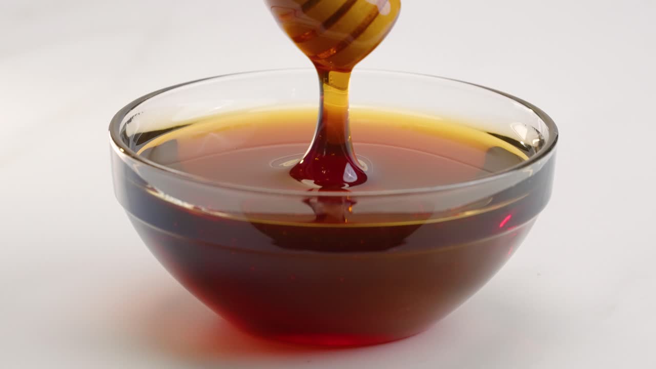 Miele mescolato in una ciotola con un bastoncino dosatore per miele, che cola e gocciola al rallentatore su sfondo bianco in 4k.