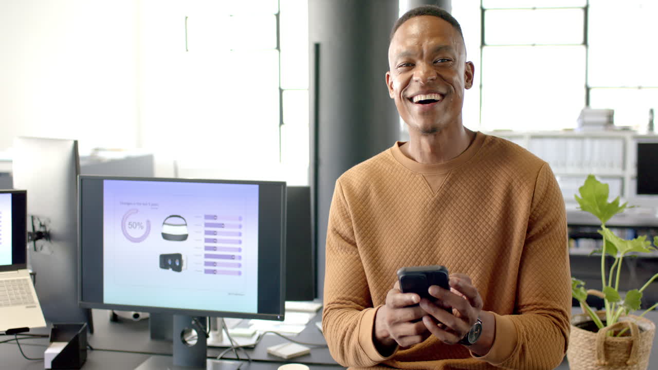 Smiling businessman holding smartphone, sitting at desk with computer in office