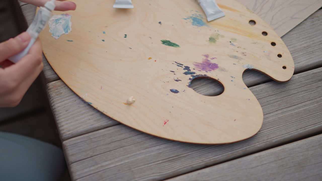 artista di strada caucasico che sistema una palette, palette ovale in legno cosparsa di macchie e sbavature di pittura, tubetti di pittura spremuti, trasferimento di colore sulla punta delle dita, panca di jeans e fogli di schizzi nelle vicinanze
