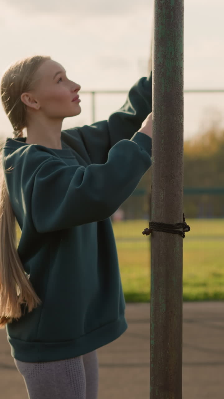 Visuale laterale di una donna che tiene una rete da pallavolo mentre altre due donne giocano a pallavolo sullo sfondo, in un contesto di sport all'aperto con cielo limpido e azione in lontananza