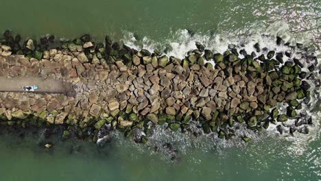 Aerial-top-down-rocky-jetty-surrounded-by-green-ocean-during-sunny-day-in-Murrells-Inlet,United-States-America