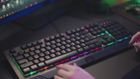 Hand view of lady in fluffy sweater adjusting RGB mechanical keyboard on desk while seated in front of monitor, fingers with polished nails repositioning keyboard under colorful ambient lighting