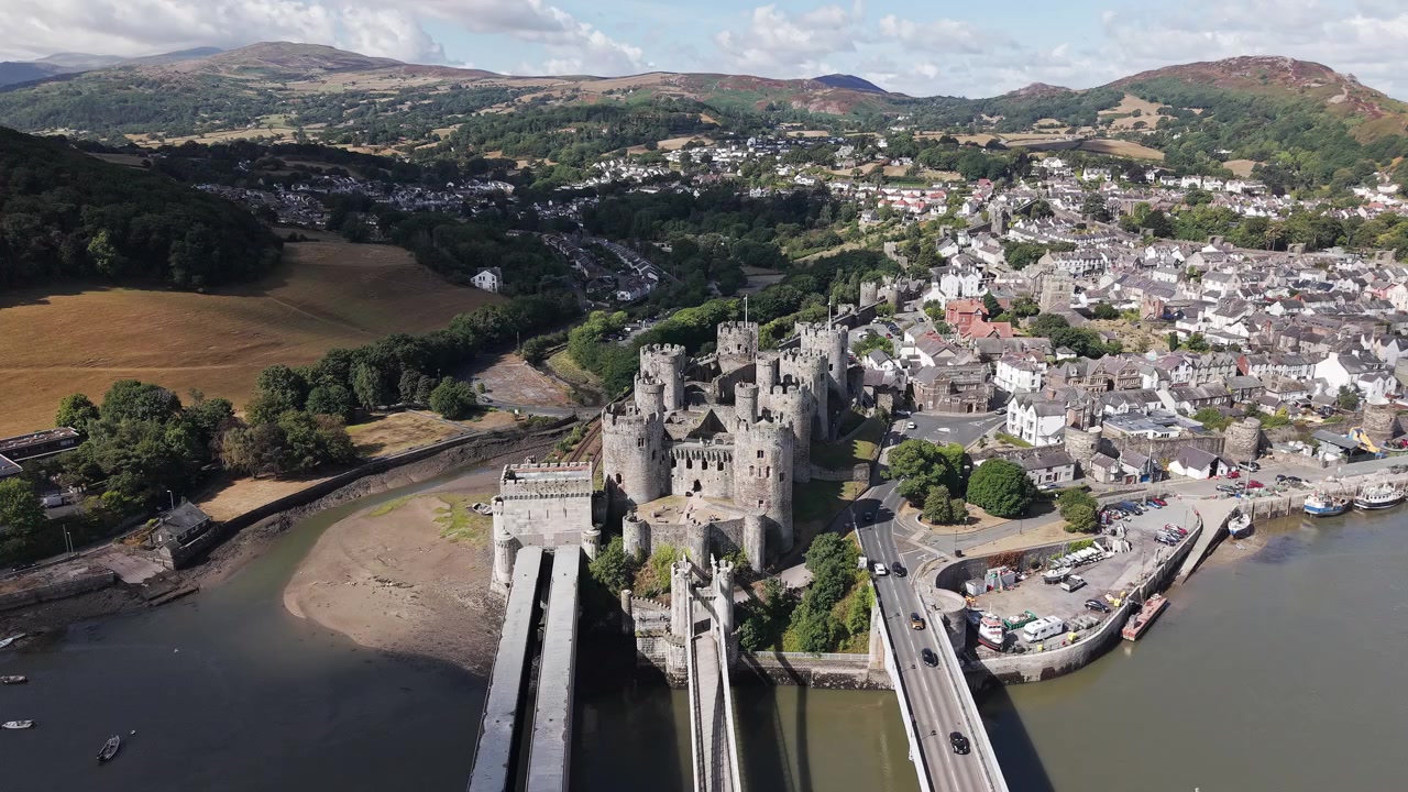 Drone pushing in over water toward ancient Conwy Castle, bridges and surrounding town, high angle aerial perspective