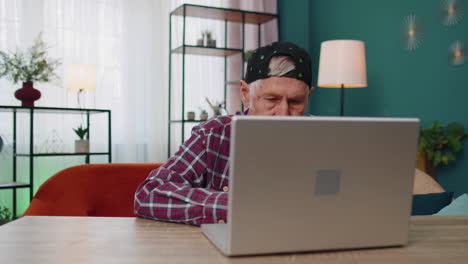 anciano abuelo hombre usando computadora portátil, escribiendo en la habitación de casa, mirando a la cámara, sonriendo