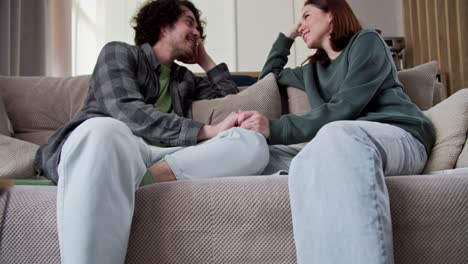 Zoom-In-bottom-view-of-a-happy-guy-brought-with-curly-hair-holding-hands-with-his-girlfriend-and-actively-laughing-while-talking-while-sitting-on-the-couch-at-home