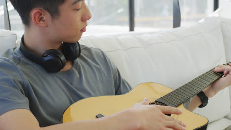 asian male teenager with headphones playing guitar and sitting in living room