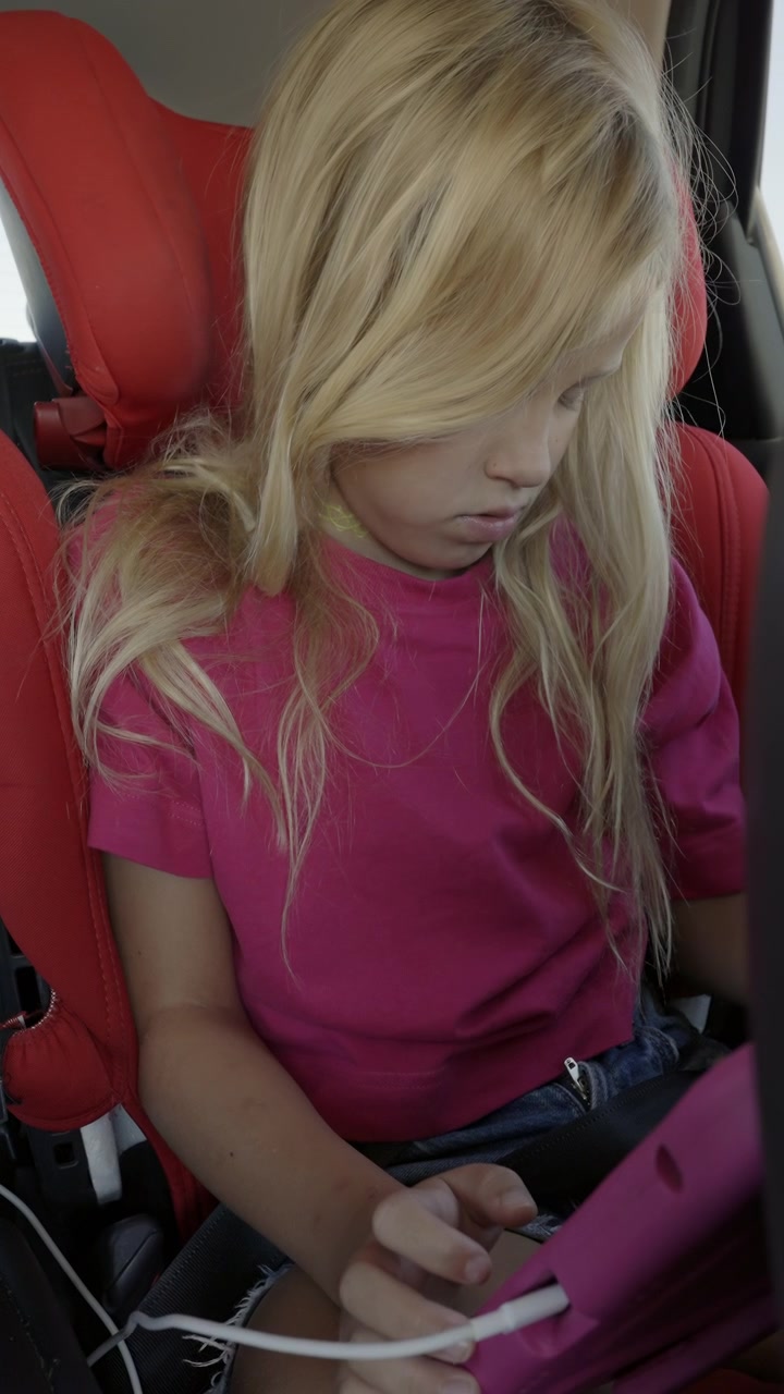 Un niño con cabello rubio largo lleva una camiseta rosa, sentado en un asiento de coche mientras se concentra en una tableta, mostrando una postura relajada y absorto en la pantalla