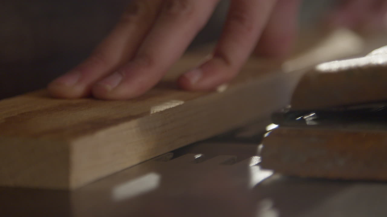 High quality video of a hand guiding a wooden board across a saw in warm cinematic light. Slow motion captures dust, grain and precise woodworking in a calm workshop atmosphere