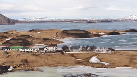 aerial view of reykjahlíð village and volcanic pseudo-craters on the shores of partially frozen lake mývatn, iceland, with distant mountains.